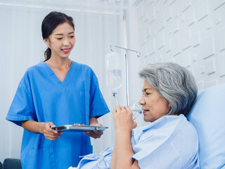 Asian elderly patient taking medicine, hold drinking water glass and pill from young nurse in blue scrub suit in hospital room, daily medicine or vitamin supplements, senior healthcare and medical.