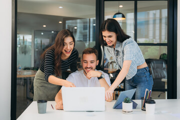 Diverse, ecstatic businesspeople discussing papers and working on laptops at an office table while smiling.