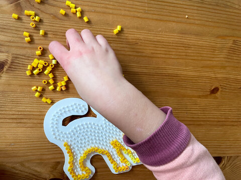 Top down Aufnahe auf eine Kinderhand, die gelbe B&uuml;gelperlen auf ein Motiv in Katzenform steckt, als Zeitvertreib im Herbst oder WInter 