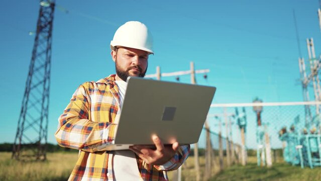 Design engineer in helmet holds laptop in hands at construction site in field, estimating investment attractiveness this object. Male compares his project with customer project. Using technologies.