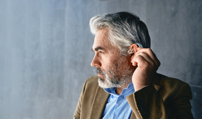 Grey-haired mature man with hearing impairment using hearing aid. Hearing solutions for deafness people