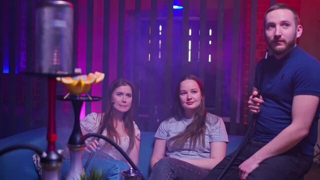 two young women and a young man of European origin are sitting in a hookah lounge, chatting, laughing, smiling and smoking a hookah