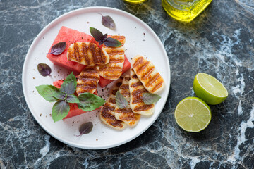 White plate with watermelon, grilled haloumi cheese and fresh basil, elevated view on a black marble background, horizontal shot