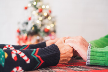 Senior couple in warm clothing holding each others hands in front of decorated christmas tree at home. Loving old romantic heterosexual couple celebrating christmas festival together.