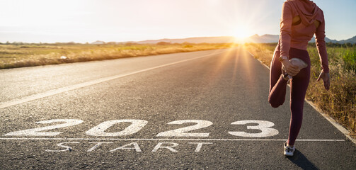 New year 2023 or start straight concept.word 2023 written on the asphalt road and athlete woman runner stretching leg preparing for new year at sunset.Concept of challenge or career path and change. - obrazy, fototapety, plakaty
