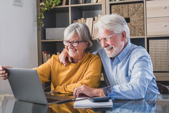 Happy elderly 60s couple sit rest on couch at home pay household expenses online on computer, smiling mature 50s husband and wife clients hold documents make payment on internet banking service.