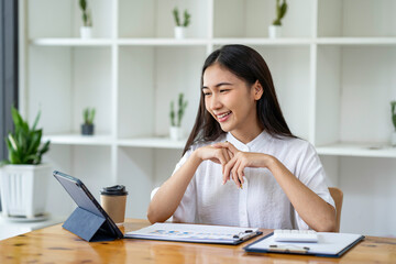 An Asian businesswoman wipes a to-do list on a tablet and responds to customer chats at a desk in a...
