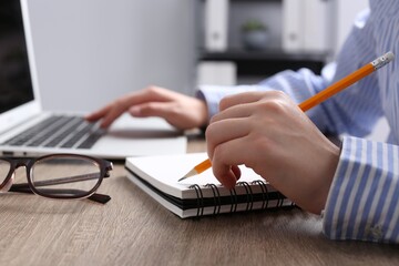 Woman with notebook working on laptop at wooden table in office, closeup