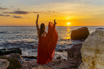 a dancing woman in a red flying dress on the ocean or on the sea beach against the backdrop of the sunset sky.