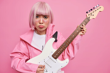 Fototapeta premium Dissatisfied Asian woman with pink dyed hair plays electic acoustic guitar learns playing musical instrument dressed in jacket frowns face has bad mood isolated over rosy background. Upset musician