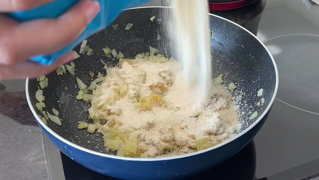 Preparing farofa (Brazilian toasted cassava flour) toasting in a pan on the electric stove. Adding the cassava flour and stirring continuously with a wooden spoon.