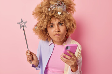 Annoyed irritated curly haired woman pouts lips looks angrily at camera holds mobile phone and magic wand sends messages of invitation on party wears festive clothes isolated over pink background