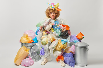Naklejka premium Shocked woman covered with plastic garbage saves environment from pollution collects plastic litter for recycling surrounded by bins stares at camera isolated over grey background. Ecology concept