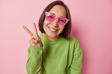 Positive Asian woman with dark hair makes peace sign enjoys amusing time wears sunglasses and green pullover smiles gladfully isolated over pink background. People and body language concept.