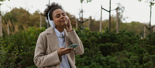 a young african american woman in a beige coat listens to music in headphones with her eyes closed against the backdrop of a city park