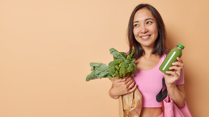 Sporty smiling Asian woman with dark hair holds paper bag of fresh vegetables and smoothie keeps to...