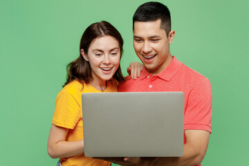 Young smiling happy cheerful fun IT couple two friends family man woman in basic t-shirts together hold use work on laptop pc computer isolated on pastel plain light green background studio portrait
