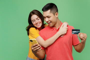 Young happy couple two friends family man woman wear t-shirts together use mobile cell phone credit bank card shopping online order delivery hugging isolated on pastel plain light green background. - obrazy, fototapety, plakaty