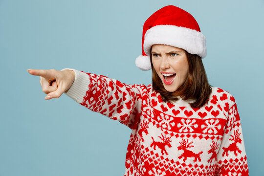 Young angry sad mad woman 20s wear warm red knitted sweater Santa hat posing point index finger aside scream isolated on plain pastel light blue cyan background. Happy New Year 2023 holiday concept.