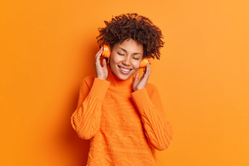 Horizontal shot of satisfied curly haired woman keeps hands on headphones keeps eyes closed from...