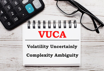 On a light wooden table calculator, glasses and a blank notepad with the text VUCA Volatility...