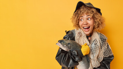 Horizontal shot of happy surprised female angler poses with fish net and stout has good successful day for fishing dressed in leather hat and raincoat isolated over yellow background blank space