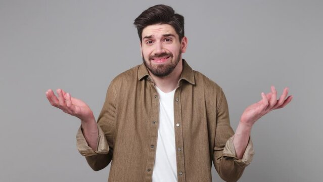 Young fun confused shy shamed man 20s he wears brown shirt look camera spreading hands say oops ouch oh omg i am so sorry isolated on plain grey background studio portrait. People lifestyle concept