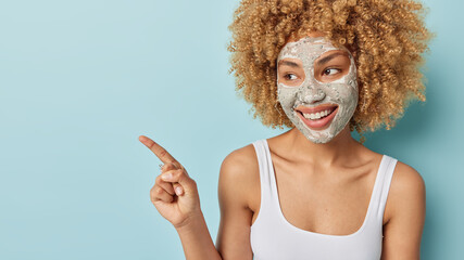 Horizontal shot of beautiful young woman with curly hair applies beauty facial mask for skin...