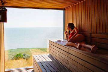 woman in a red swimsuit lies on a bench in the sauna with the panoramic window. 