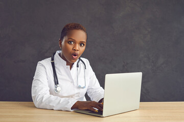 Dark skinned female doctor with shocked face sitting at the table on a gray background. Surprised...