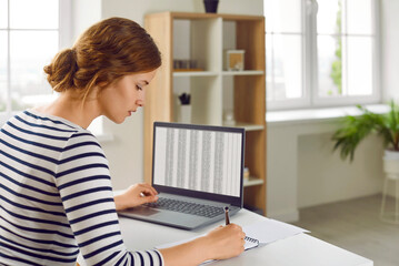 Young woman office worker is working with spreadsheet on computers screens, making notes writing in notebook. Business analytics, accounting, finance, data analytics, data science, marketing concept.