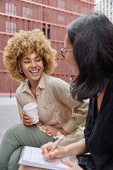 Outdoor shot of two happy diverse female students meet together discuss future project write down information in notebook drink coffee to go laugh joyfully have good mood. Cooperation concept