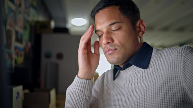 Man tired working on computer with headache at night, remove glasses and touch face in office. Exhausted latino man, take off spectacles in front of laptop or pc while working late on the internet