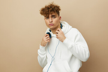 a cute, attractive man stands on a beige background in a white hoodie with headphones around his...