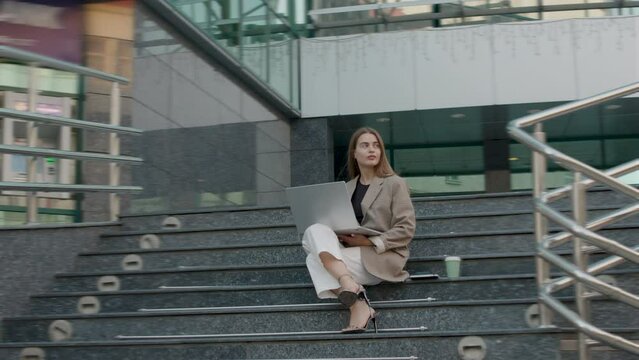Moving image of a woman dressed classically and in heels, sitting on the stairs of a modern office and working on the computer. Female director who works from anywhere