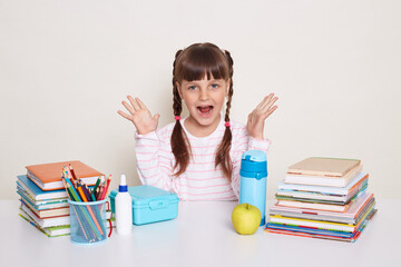 Portrait of excited positive little schoolgirl with pigtails wearing striped shirt sitting at the desk surrounded with books and other school supplies raised arms and screaming happily.