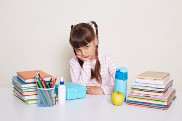 Indoor shot of sad upset little schoolgirl with dark hair and braids sitting at table surrounded with books and keeps hand under chin, being bored and exhausted, looking at camera.