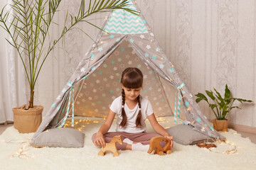 Portrait of dark haired cute little girl playing with toys and in teepee tent, sitting on floor on soft carpet, posing alone in wigwam, waiting with her friends.