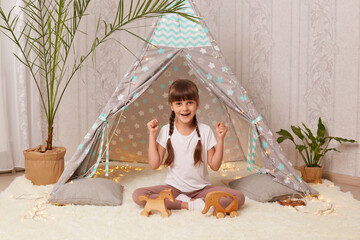 Indoor shot of extremely happy cute little girl playing in teepee tent, being in good mood, clenched fists and screaming with happiness, celebrating victory.