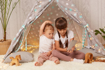 Indoor shot of adorable little girls wearing white t shirts sitting on floor in wigwam and looking at smart phone display, infant baby raised arms, elder sister holding smart phone in hands.