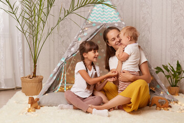 Image of smiling satisfied little girls and her mother playing at home in wigwam and being happy to spend time together, female hugging her kids, being in good mood. © sementsova321