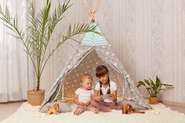 Image of c harming cute little girls wearing white t shirts sitting on floor in shelter and playing on mobile phone or watching cartoons, being in good mood, having fun together. © sementsova321