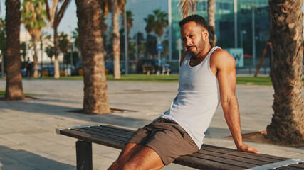 Young bearded male athlete is resting after training sitting on bench on modern background