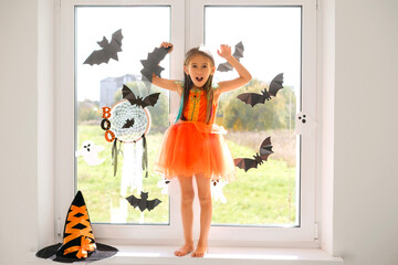 Kid stands on windowsill at the decorated window of children's room with decorations for Halloween and makes a frightening gesture with his hands. The girl is preparing the nursery for the holiday