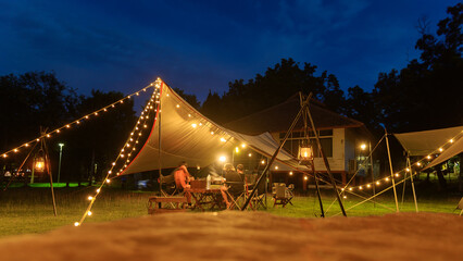 Outdoor camping tent with tarp or flysheet on grass courtyard and warm night light under dark blue...