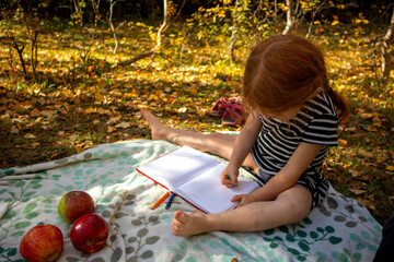 a little red-haired girl draws on a picnic in autumn on a blanket. there are apples nearby