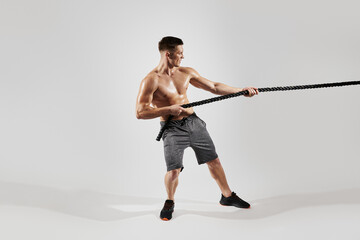 Concentrated young man with perfect body pulling a rope against white background