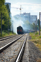 The freight train is driven by a diesel locomotive. railroad on the outskirts of the city. a lot of gray smoke escapes from the locomotive chimney