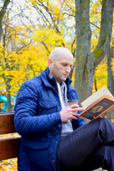 Fototapeta premium young man reads a book and drinks coffee outdoors, sitting on a bench in an autumn park. serenity, relaxation and privacy concept