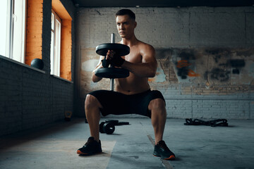 Confident young man standing in squatting position while training with dumbbell in gym